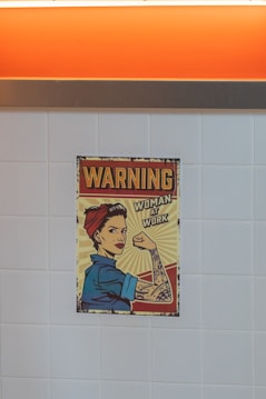 A vintage-style poster depicting a woman with a confident expression, dressed in a blue work shirt and red bandana, reminiscent of the 'Rosie the Riveter' iconography. The woman is flexing her arm to show her strength, with tattoos visible. The poster includes the text 'Warning' and 'Woman at Work', suggesting empowerment and gender equality. The background consists of a tiled wall and an orange panel at the top.