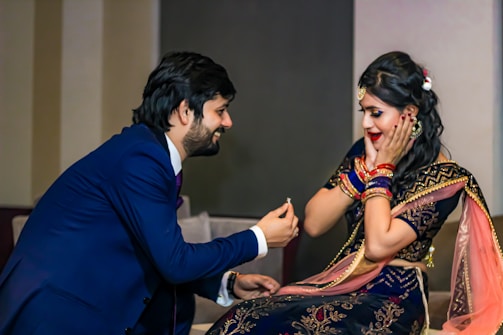 A man is kneeling with a smile while offering an engagement ring to a woman. The woman, dressed in a traditional outfit with intricate designs, appears surprised and joyful, covering her cheeks with her hands.