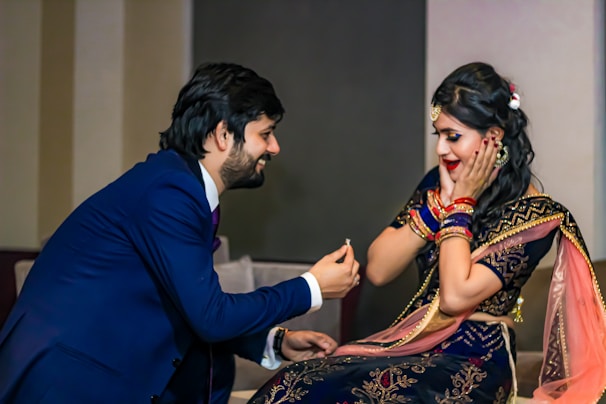 A man is kneeling with a smile while offering an engagement ring to a woman. The woman, dressed in a traditional outfit with intricate designs, appears surprised and joyful, covering her cheeks with her hands.