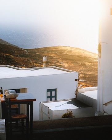 White buildings with flat roofs are perched on a hillside overlooking the ocean in the distance. A table with chairs is positioned in the foreground. The sunlight creates a warm, glowing effect over the landscape, highlighting the natural terrain and the distant sea.