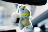 A light green plush toy dinosaur with a yellow snout and eyes, hanging from a rearview mirror beside a fluffy black and white sushi keychain. The plush dinosaur has a friendly appearance with a round rosy cheek and small yellow spikes on its head. The background suggests the interior of a car with blurred elements in the vicinity.