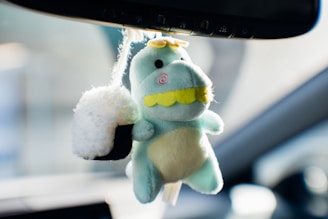 A light green plush toy dinosaur with a yellow snout and eyes, hanging from a rearview mirror beside a fluffy black and white sushi keychain. The plush dinosaur has a friendly appearance with a round rosy cheek and small yellow spikes on its head. The background suggests the interior of a car with blurred elements in the vicinity.