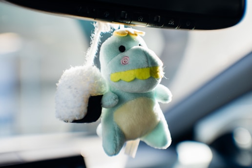 A light green plush toy dinosaur with a yellow snout and eyes, hanging from a rearview mirror beside a fluffy black and white sushi keychain. The plush dinosaur has a friendly appearance with a round rosy cheek and small yellow spikes on its head. The background suggests the interior of a car with blurred elements in the vicinity.