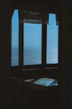 A serene view of the ocean can be seen through a large window adorned with decorative curtains. The light blue sea appears calm, creating a peaceful atmosphere. A small table and chair are visible in the foreground, framed by the silhouette of the room's interior.