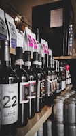 A selection of various wine bottles and canned drinks neatly arranged on wooden shelves. Wine bottles display different labels, some with visible years and origins, while cans are positioned at the edge, with soft drink or beer labels. Above the drinks, coffee packages in neat rows show colorful labels and designs. Lighting creates a sophisticated and inviting ambiance in the setting.