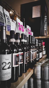A selection of various wine bottles and canned drinks neatly arranged on wooden shelves. Wine bottles display different labels, some with visible years and origins, while cans are positioned at the edge, with soft drink or beer labels. Above the drinks, coffee packages in neat rows show colorful labels and designs. Lighting creates a sophisticated and inviting ambiance in the setting.