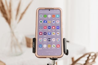 A smartphone is mounted on a tripod, showcasing a variety of app icons on its screen. The background is softly focused, revealing hints of an indoor setting with dried plants and furniture.