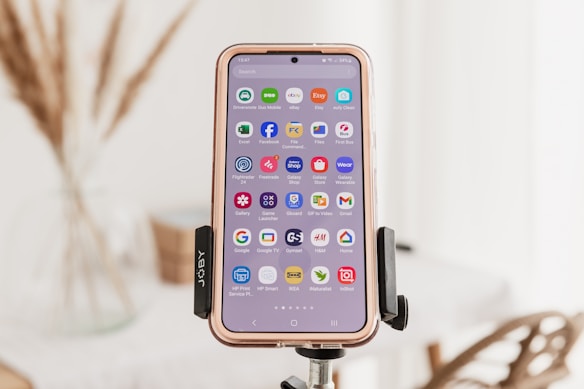A smartphone is mounted on a tripod, showcasing a variety of app icons on its screen. The background is softly focused, revealing hints of an indoor setting with dried plants and furniture.