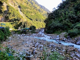 A serene mountain landscape features a flowing river surrounded by lush green hills. Rocky terrain lines the riverbanks, and a small structure is visible on the left side near the forested area.