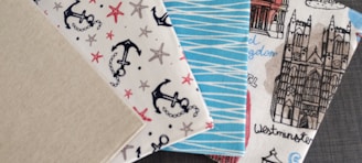 Four patterned fabric swatches are displayed. The first swatch has a plain beige color. The second one is embellished with nautical-themed designs featuring black anchors and red and brown starfish on a white background. The third swatch displays a blue and white zigzag pattern. The fourth swatch illustrates architectural elements, including a detailed drawing of a gothic-style cathedral with handwritten labels such as 'Westminster'.