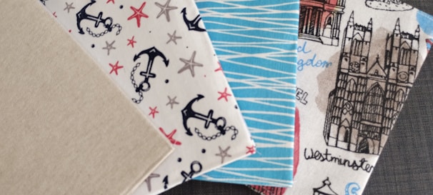 Four patterned fabric swatches are displayed. The first swatch has a plain beige color. The second one is embellished with nautical-themed designs featuring black anchors and red and brown starfish on a white background. The third swatch displays a blue and white zigzag pattern. The fourth swatch illustrates architectural elements, including a detailed drawing of a gothic-style cathedral with handwritten labels such as 'Westminster'.