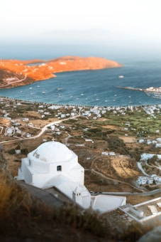 A white-domed building typical of Greek architecture is positioned in the foreground, overlooking a scenic coastal village. The landscape includes several white buildings scattered across brown and green fields. A deep blue sea stretches beyond, dotted with numerous sailboats. The distant hills appear golden under the sunlight, contrasting with the clear sky above.