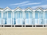 A row of pastel blue and white striped beach huts stands in a neat formation. Each hut has a peaked roof and a small porch, with shadows cast on the ground. The sandy beach stretches out in front, and the sky above is clear with a few wispy clouds.
