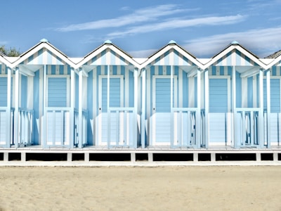 A row of pastel blue and white striped beach huts stands in a neat formation. Each hut has a peaked roof and a small porch, with shadows cast on the ground. The sandy beach stretches out in front, and the sky above is clear with a few wispy clouds.