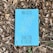 A book with a bright blue cover featuring the title 'TRADUTOR DE CHUVAS' and the author's name, Mia Couto, is placed on a bed of dry, brown leaves. The cover includes abstract yellow and white linear designs.