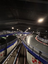 An indoor go-kart track with elevated sections and multiple curves, featuring blue barriers and overhead lights. The environment is industrial, with a dimly lit atmosphere.