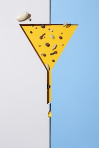 A geometric composition features a yellow funnel shape on a split background of white and blue. Various spices, including cardamom, cloves, and peppercorns, are arranged inside the yellow funnel. A single drop of liquid falls from the narrow end of the funnel.