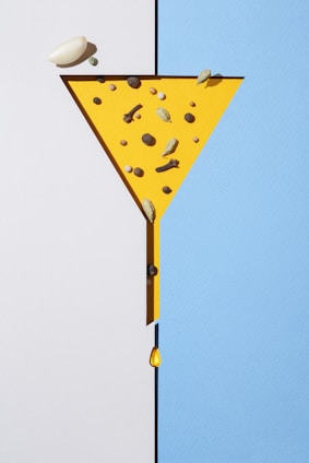 A geometric composition features a yellow funnel shape on a split background of white and blue. Various spices, including cardamom, cloves, and peppercorns, are arranged inside the yellow funnel. A single drop of liquid falls from the narrow end of the funnel.