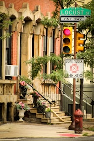 A street corner with a red brick building featuring ornate window detailing, a staircase with wrought iron railings, and potted plants with colorful flowers. A Locust Street sign, one-way sign, a traffic light with a red light, and a 'Don't Block the Box' sign are visible. A red fire hydrant is situated on the sidewalk near the corner.
