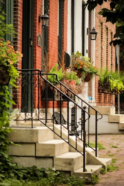 A row of charming, brick townhouses features black metal railings, small staircases leading up to the front doors, and window boxes filled with a variety of lush green plants and colorful flowers. Black lantern-style wall lights are mounted next to each door, and the ground is paved with cobblestones. A few overhanging trees and a variety of greenery add a natural touch to the scene.