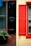 A vibrant contrast between two window shutters, one painted black on a brick wall and the other red on a yellow-painted wall. Below the black shutter, a planter box holds green foliage and bright red flowers. Adjacent windows feature drawn blinds.