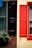 A vibrant contrast between two window shutters, one painted black on a brick wall and the other red on a yellow-painted wall. Below the black shutter, a planter box holds green foliage and bright red flowers. Adjacent windows feature drawn blinds.