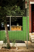 The image features a storefront with a green tiled exterior wall and a window displaying a humorous sign that reads 'DROP YOUR PANTS OFF TO US'. A tree with green leaves is in front, along with a sidewalk and a 'NO STOPPING ANY TIME' street sign.