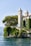 A picturesque villa with two tall, elegant towers located on a lush, green cliffside overlooking a serene body of water. The villa is partially covered in climbing vines, adding to its charm. Mature trees surround the property, providing shade and natural beauty. The sky is clear, contributing to the tranquil ambiance.