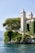 A picturesque villa with two tall, elegant towers located on a lush, green cliffside overlooking a serene body of water. The villa is partially covered in climbing vines, adding to its charm. Mature trees surround the property, providing shade and natural beauty. The sky is clear, contributing to the tranquil ambiance.