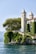 A picturesque villa with two tall, elegant towers located on a lush, green cliffside overlooking a serene body of water. The villa is partially covered in climbing vines, adding to its charm. Mature trees surround the property, providing shade and natural beauty. The sky is clear, contributing to the tranquil ambiance.