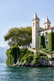 A picturesque villa with two tall, elegant towers located on a lush, green cliffside overlooking a serene body of water. The villa is partially covered in climbing vines, adding to its charm. Mature trees surround the property, providing shade and natural beauty. The sky is clear, contributing to the tranquil ambiance.