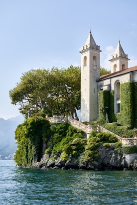 A picturesque villa with two tall, elegant towers located on a lush, green cliffside overlooking a serene body of water. The villa is partially covered in climbing vines, adding to its charm. Mature trees surround the property, providing shade and natural beauty. The sky is clear, contributing to the tranquil ambiance.