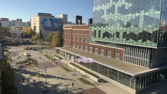 The image captures a modern urban setting with a mix of architectural styles. A large, contemporary glass building features geometric patterns on its facade, and an adjacent brick building adds a classic touch. In the distance, a mural can be seen on a building, adding vibrant colors to the scene. The area appears to be a courtyard or plaza, with walkways, trees, and several pedestrians moving about. The setting is bright and sunny, indicating a clear day.