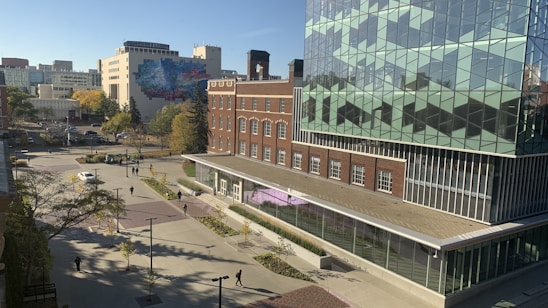 The image captures a modern urban setting with a mix of architectural styles. A large, contemporary glass building features geometric patterns on its facade, and an adjacent brick building adds a classic touch. In the distance, a mural can be seen on a building, adding vibrant colors to the scene. The area appears to be a courtyard or plaza, with walkways, trees, and several pedestrians moving about. The setting is bright and sunny, indicating a clear day.