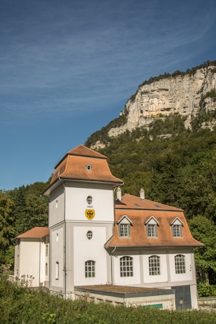 A historic building with a light gray facade and a reddish-brown roof stands in the foreground. The building features arched windows and a crest with the year 1882. Behind it, a lush green forest rises to meet a rocky cliff under a clear blue sky.