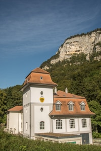 A historic building with a light gray facade and a reddish-brown roof stands in the foreground. The building features arched windows and a crest with the year 1882. Behind it, a lush green forest rises to meet a rocky cliff under a clear blue sky.