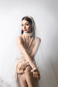 A woman sits gracefully wearing a lace dress and adorned with large pearl jewelry including bracelets, necklace, and headband. A delicate veil drapes over her, complementing the elegant look.