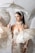 A woman is elegantly dressed in a lace gown adorned with pearls. She is surrounded by intricately designed lace parasols and has a pearl necklace and headband. Her hair is neatly styled, and she is looking to the side with a composed expression.