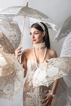A woman is elegantly dressed in a lace gown adorned with pearls. She is surrounded by intricately designed lace parasols and has a pearl necklace and headband. Her hair is neatly styled, and she is looking to the side with a composed expression.