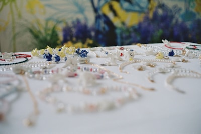 A collection of colorful jewelry, including necklaces and bracelets, is spread across a white surface. The pieces are adorned with various beads and charms in shades of blue, yellow, red, and white. The background features a blurred floral pattern with dominant blue and green hues.