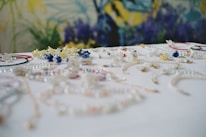A collection of colorful jewelry, including necklaces and bracelets, is spread across a white surface. The pieces are adorned with various beads and charms in shades of blue, yellow, red, and white. The background features a blurred floral pattern with dominant blue and green hues.