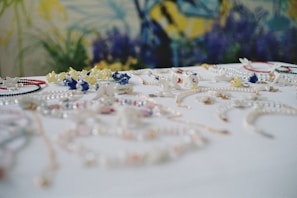 A collection of colorful jewelry, including necklaces and bracelets, is spread across a white surface. The pieces are adorned with various beads and charms in shades of blue, yellow, red, and white. The background features a blurred floral pattern with dominant blue and green hues.
