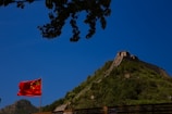 A section of the Great Wall of China stretches along a lush green hillside under a clear blue sky. A Chinese flag waves prominently in the foreground, while tree branches frame the top of the image.