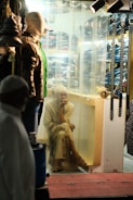A seated person is gazing outward through a clear curtain in a dimly lit clothing store. Mannequins displaying various attire, including hats and jackets, are positioned nearby, and a row of belts hangs on the right. Behind the curtain, shelves are stacked with folded clothes.
