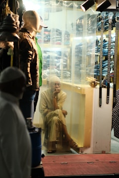 A seated person is gazing outward through a clear curtain in a dimly lit clothing store. Mannequins displaying various attire, including hats and jackets, are positioned nearby, and a row of belts hangs on the right. Behind the curtain, shelves are stacked with folded clothes.