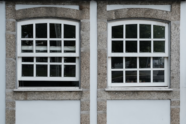 Two adjacent windows with white frames and multiple panes are set in a stone wall. Each window features a row of square panes at the top and rectangular panes below. The wall around the windows is constructed from textured gray stone with white borders. Different shades of green from trees are visible through the glass panes.