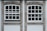 Two adjacent windows with white frames and multiple panes are set in a stone wall. Each window features a row of square panes at the top and rectangular panes below. The wall around the windows is constructed from textured gray stone with white borders. Different shades of green from trees are visible through the glass panes.