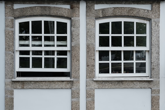 Two adjacent windows with white frames and multiple panes are set in a stone wall. Each window features a row of square panes at the top and rectangular panes below. The wall around the windows is constructed from textured gray stone with white borders. Different shades of green from trees are visible through the glass panes.