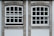 Two adjacent windows with white frames and multiple panes are set in a stone wall. Each window features a row of square panes at the top and rectangular panes below. The wall around the windows is constructed from textured gray stone with white borders. Different shades of green from trees are visible through the glass panes.