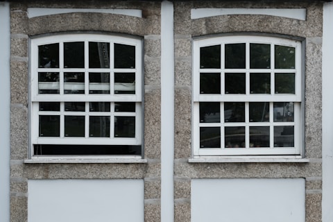 Two adjacent windows with white frames and multiple panes are set in a stone wall. Each window features a row of square panes at the top and rectangular panes below. The wall around the windows is constructed from textured gray stone with white borders. Different shades of green from trees are visible through the glass panes.